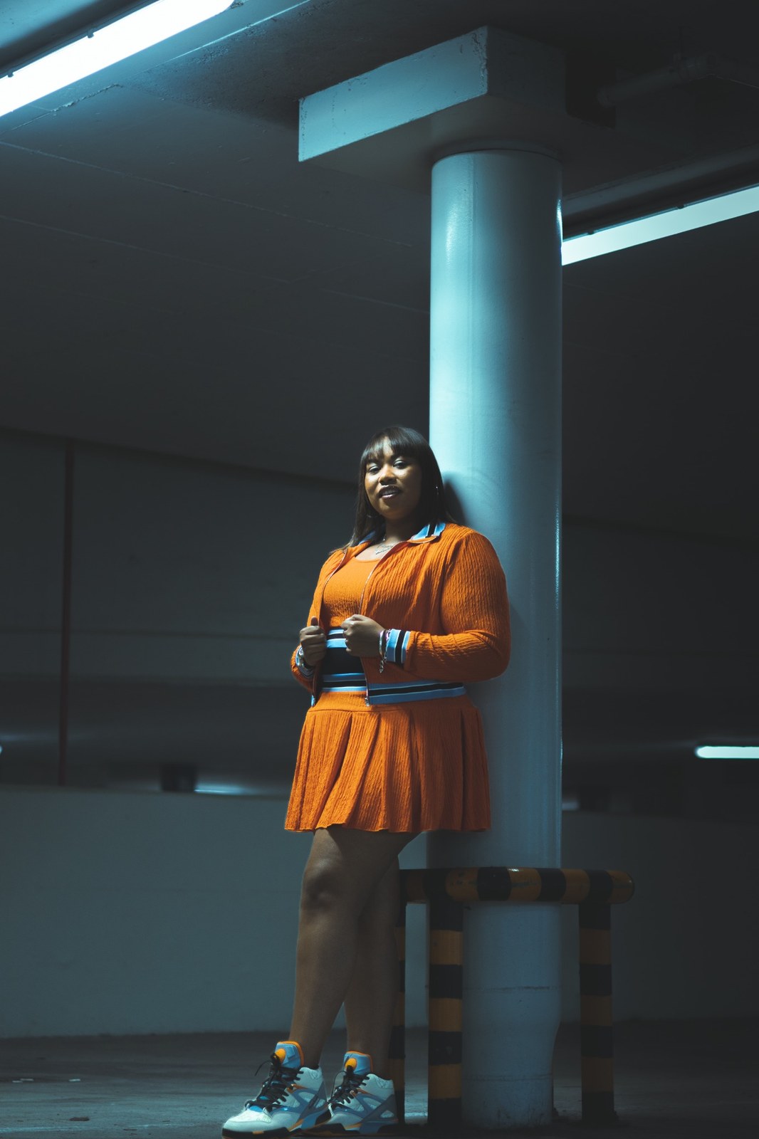 Woman in an orange outfit beside a column under blue parking-garage light