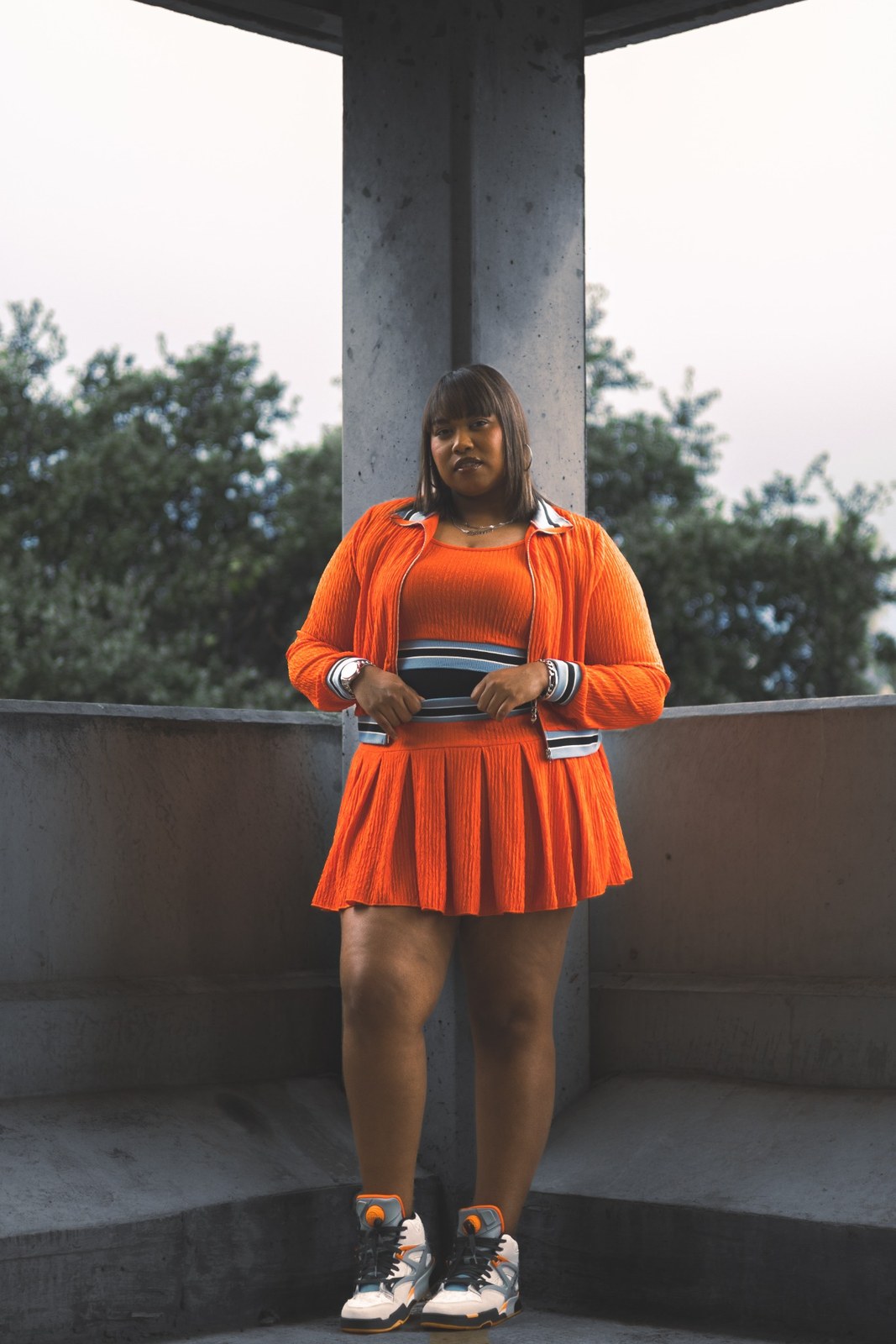 Woman standing between concrete pillars in an orange skirt and trainers